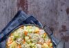 Chicken and Dumplings A cast iron pot filled with thick chicken and dumplings stew, featuring large herb dumplings, chunks of chicken, diced carrots, celery, and potatoes, seasoned with thyme, set on a dark wooden surface with a blue cloth and antique spoons.