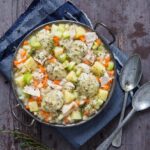 A cast iron pot filled with thick chicken and dumplings stew, featuring large herb dumplings, chunks of chicken, diced carrots, celery, and potatoes, seasoned with thyme, set on a dark wooden surface with a blue cloth and antique spoons.