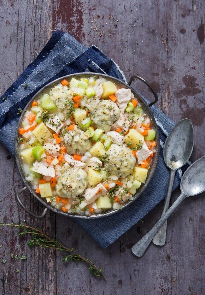 A cast iron pot filled with thick chicken and dumplings stew, featuring large herb dumplings, chunks of chicken, diced carrots, celery, and potatoes, seasoned with thyme, set on a dark wooden surface with a blue cloth and antique spoons.