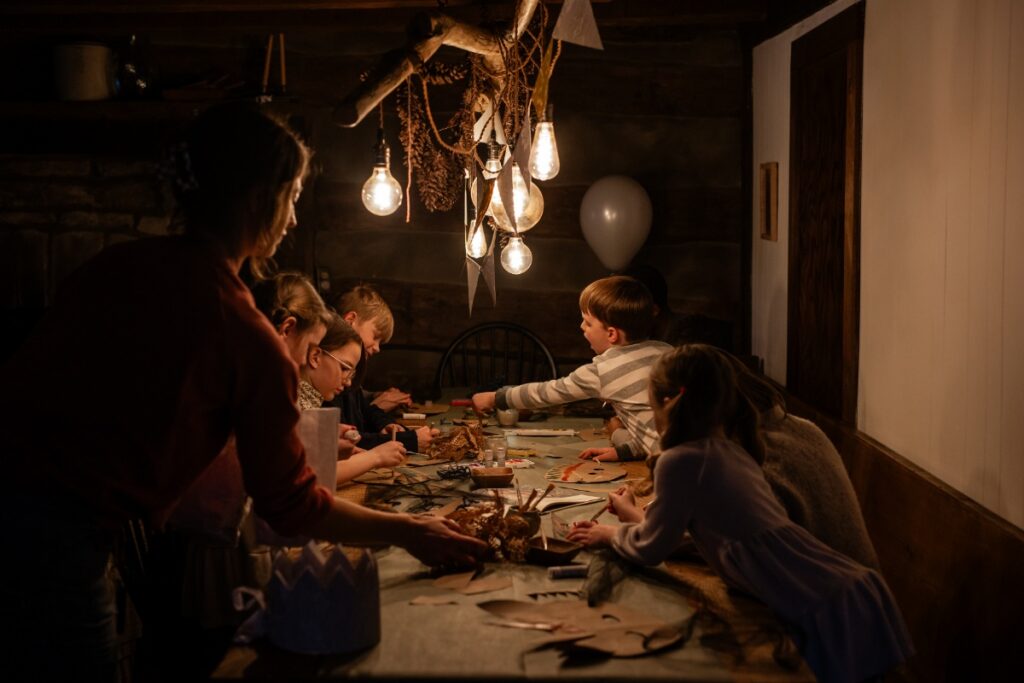 A dark, cozy interior shot of children gathered around a rustic table, crafting and decorating "Wild Thing" paper masks under hanging exposed bulb lights.
