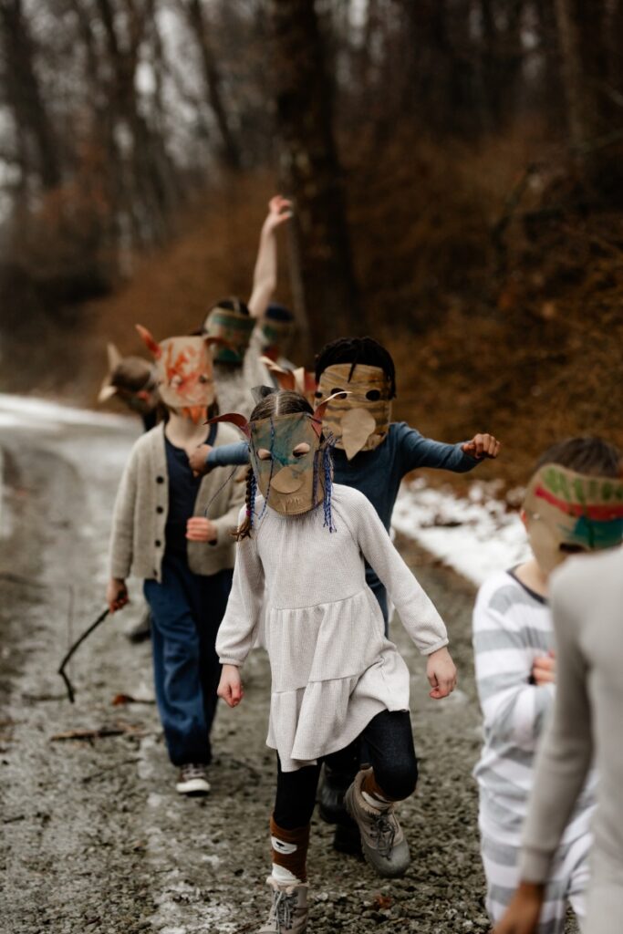 A group of children wearing elaborate, decorated paper "Wild Thing" masks run down a muddy, snowy path in a winter forest, raising their arms in a "Wild Rumpus."
