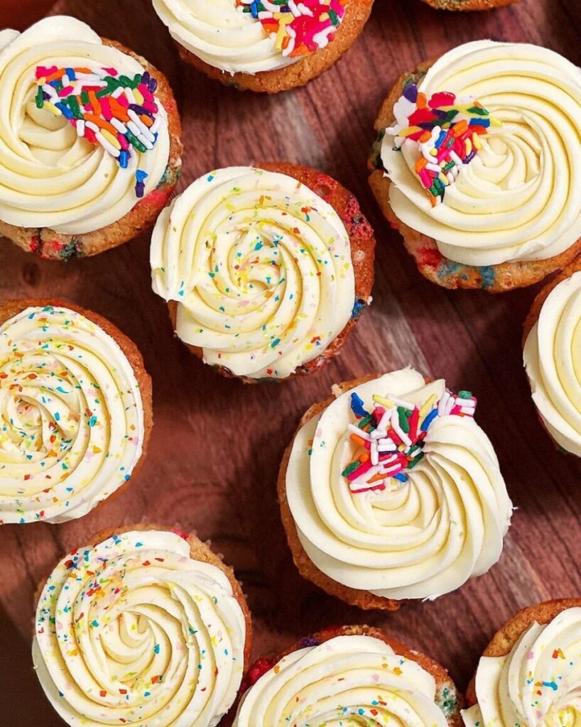Several colorful confetti cupcakes with tall swirls of white vanilla frosting and bright rainbow sprinkles, representing desserts.