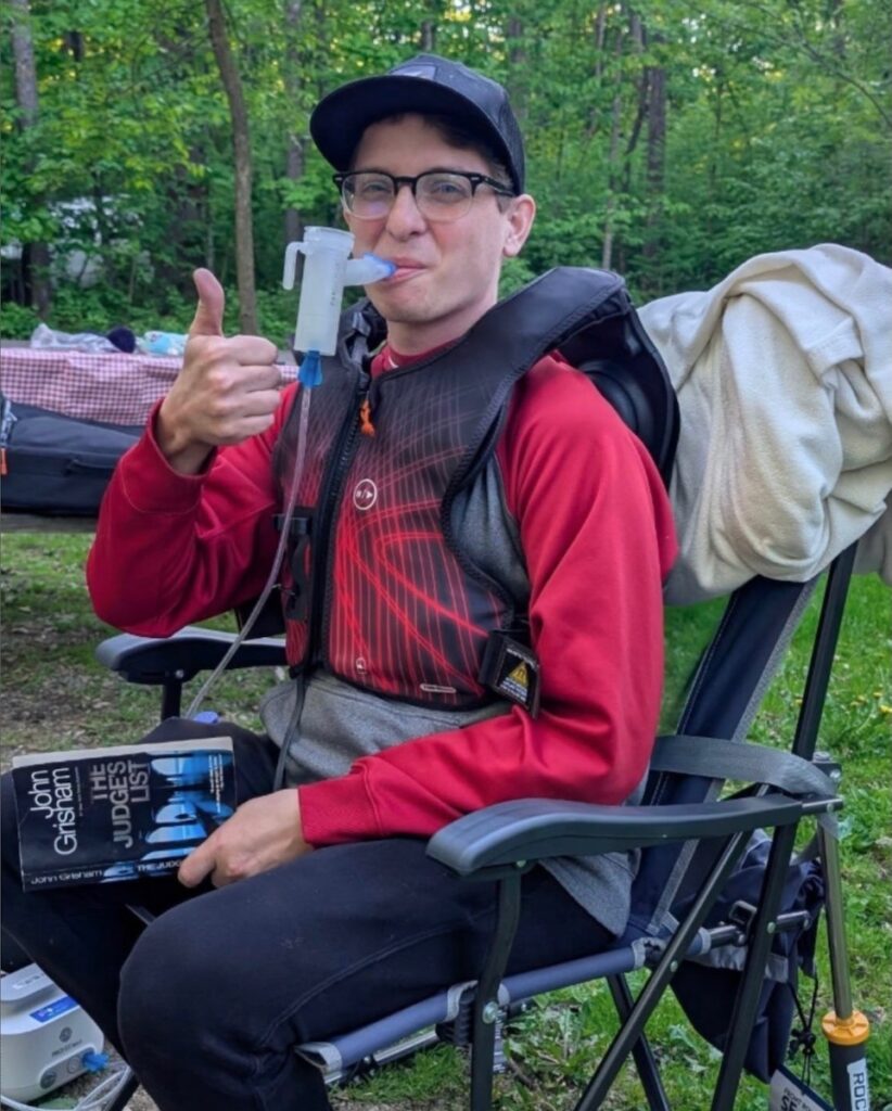 A man in a red shirt sits in a wheelchair with a breathing machine in his mouth.
