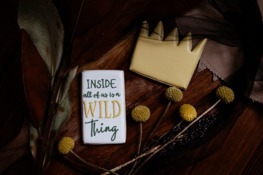 Two decorated sugar cookies, one with a yellow crown design and one with the text "INSIDE all of us is a WILD Thing," on a dark wood surface with yellow dried floral accents.