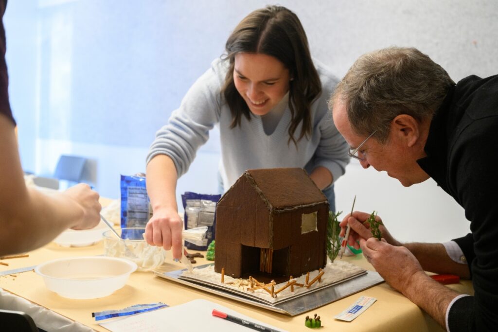 A young woman and an older man decorating a dark brown gingerbread house with white icing and green rosemary, collaboratively working on a gingerbread competition entry.