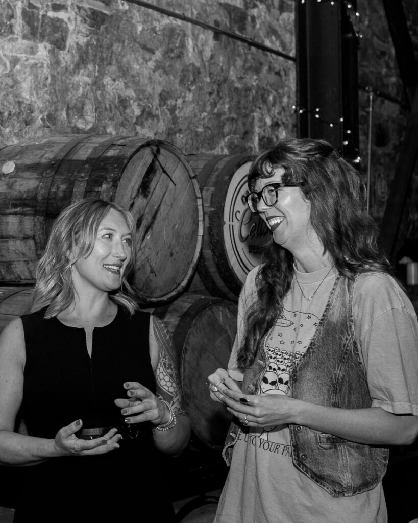 A black and white photo of Emily Fitzgerald and a friend smiling and talking, standing in front of stacked wooden barrels at a non-profit mixer or social event.