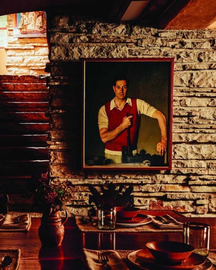 Excursion at Fallingwater TABLE Magazine A framed portrait of Edgar Kaufmann Sr. in a red vest and white shirt, holding a walking stick, hanging on a textured stone wall above the set dining table at Frank Lloyd Wright's Fallingwater.