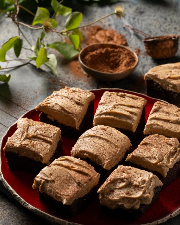 Extra Dark Brownies with Coffee Mascarpone Frosting TABLE Magazine Nine extra dark brownies topped with a swirl of coffee mascarpone frosting and a dusting of cocoa powder, arranged on a red ceramic plate for dessert.