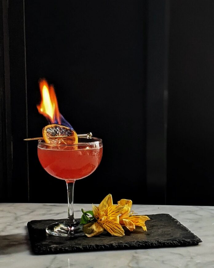 A red cocktail in a coupe glass with a flaming, rum-soaked citrus garnish, sitting on a black slate tray next to a yellow flower, against a dark background.