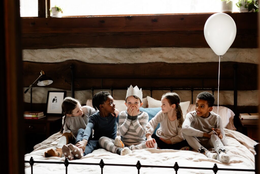 A birthday party image of five children in pajamas sitting on a bed, smiling, with one boy in a paper crown playfully covering his mouth, and a white balloon floating next to the bedpost.