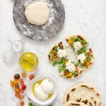 Fresh flatbread pizza topped with burrata cheese, arugula, and cherry tomatoes, alongside a ball of dough on a wooden board, olive oil, and a bowl of fresh burrata on a marble countertop.