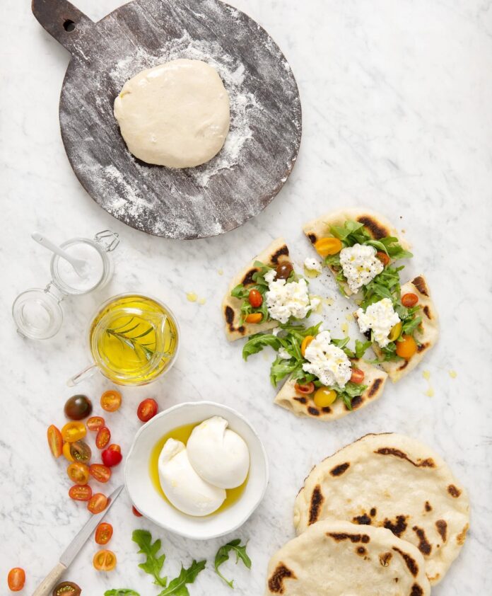 Fresh flatbread pizza topped with burrata cheese, arugula, and cherry tomatoes, alongside a ball of dough on a wooden board, olive oil, and a bowl of fresh burrata on a marble countertop.