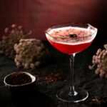 A deep pink Floral Fumeé cocktail in a coupe glass with a gold rim and foam, garnished with a star anise and hibiscus flakes, surrounded by dried flowers and spices.