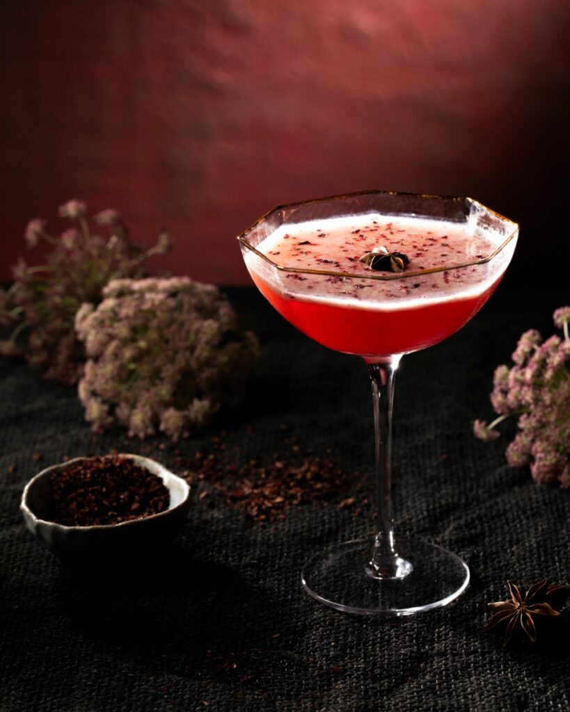 A deep pink Floral Fumeé cocktail in a coupe glass with a gold rim and foam, garnished with a star anise and hibiscus flakes, surrounded by dried flowers and spices.