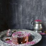 A molten chocolate lava cake dusted with powdered sugar and sprinkled with bright pink salt, revealing a gooey center, served on a distressed metal plate next to a silver sugar pitcher.
