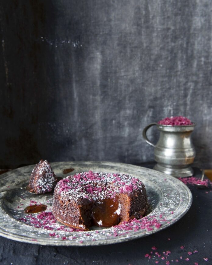 A molten chocolate lava cake dusted with powdered sugar and sprinkled with bright pink salt, revealing a gooey center, served on a distressed metal plate next to a silver sugar pitcher.