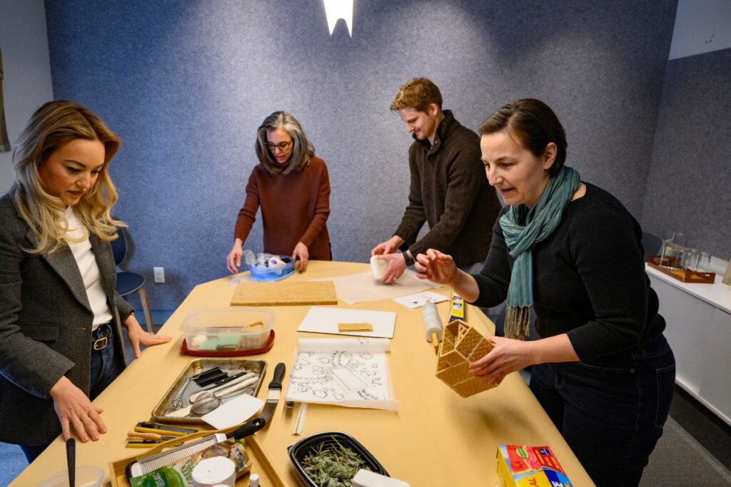 Four architects are gathered around a table, collaborating on assembling and decorating a gingerbread house during an office holiday competition.