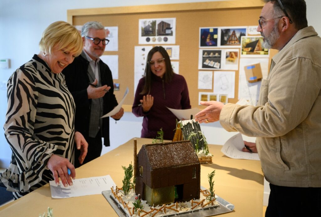 Four smiling judges and architects are gathered around a table with two modern gingerbread houses, discussing the competition during the pitch presentations.