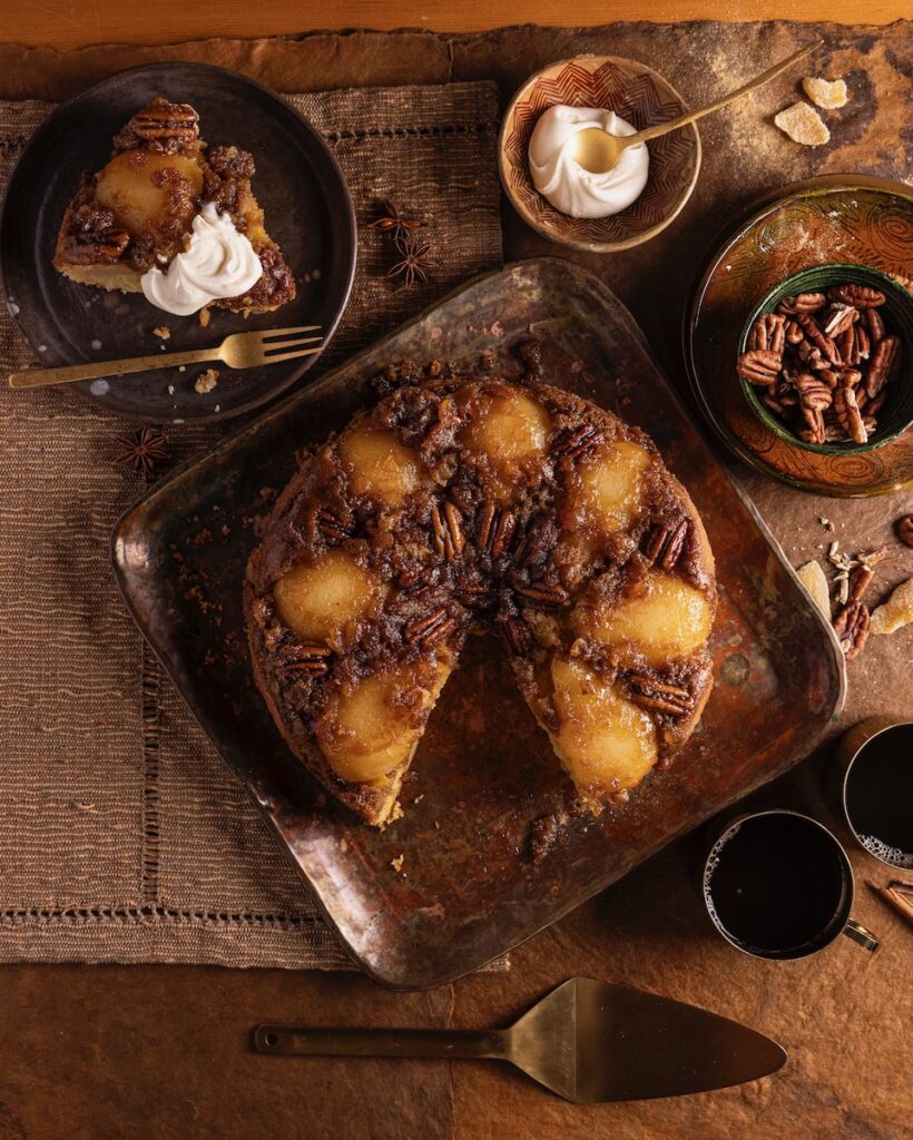 A pear upside down cake sits on a wooden table with a slice taken out of it on a plate to the top left.