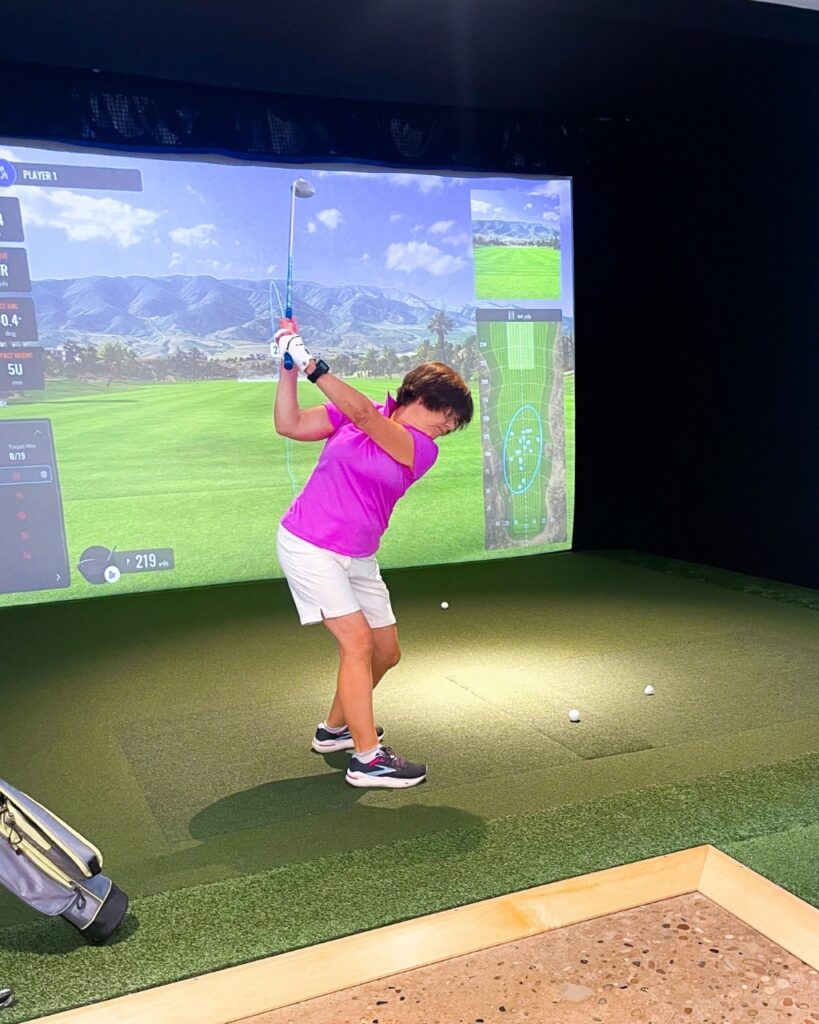 A woman in a pink shirt golfing indoors to a screen.