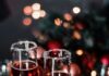 The Best Wines for Your Holiday Dinner Table Two champagne flutes with red wine on a holiday table with lights and bulbs.