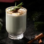 A Holiday in San Juan coquito cocktail, garnished with a rosemary sprig, cinnamon stick, and pistachio-dusted rim, set on a dark tablecloth with pine needles.