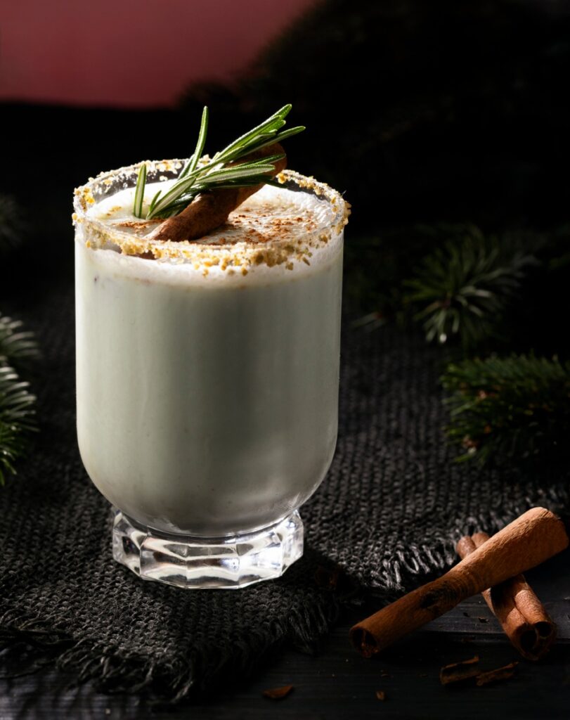 A Holiday in San Juan coquito cocktail, garnished with a rosemary sprig, cinnamon stick, and pistachio-dusted rim, set on a dark tablecloth with pine needles.