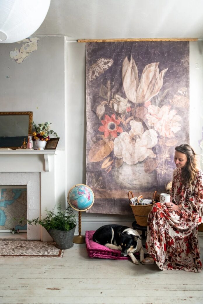 KonMari Method Home Design A woman, Quelcy Kogel, in a long floral dress sits with a coffee cup next to a large, faded floral tapestry and a sleeping black and white dog on a magenta cushion in a rustic, minimalist living room.