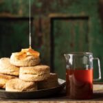 A drizzle of hot honey gets poured onto a stack of homemade buttermilk biscuits sitting beside a pitcher of hot honey.