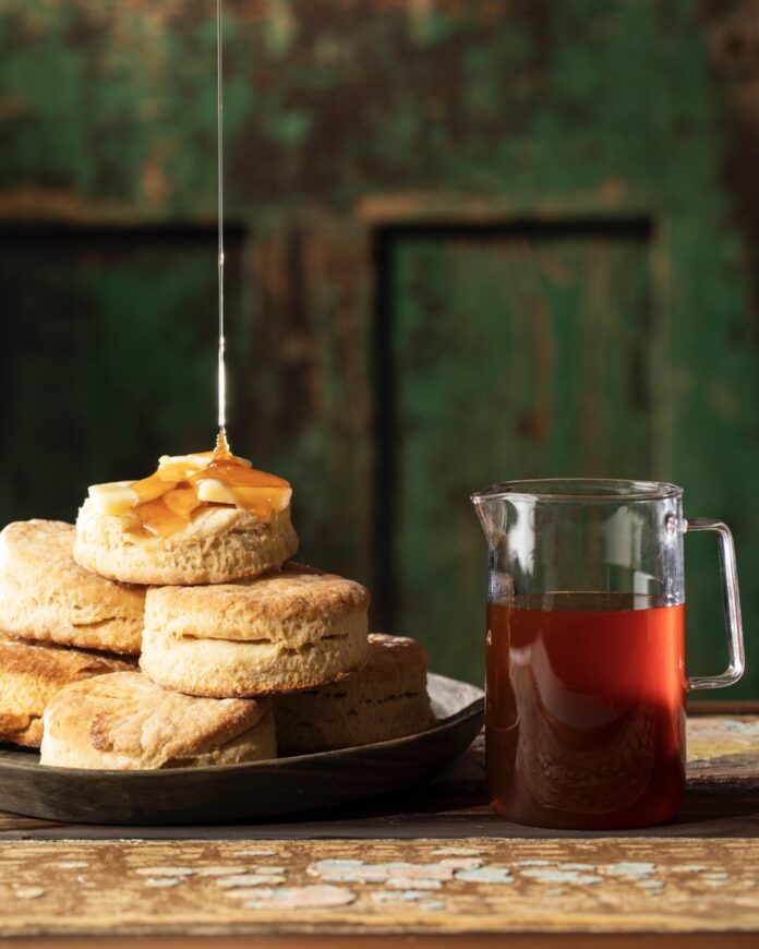 Hot Honey Buttermilk Biscuits TABLE Magazine A drizzle of hot honey gets poured onto a stack of homemade buttermilk biscuits sitting beside a pitcher of hot honey.