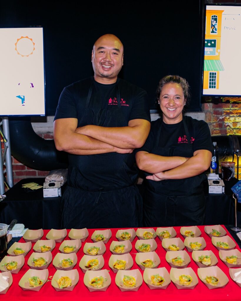 A man in a black apron with his arms crossed smiles beside a woman in a black apron with her arms crossed.