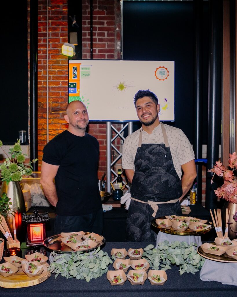 Two men chefs stand behind a table of their food samples.