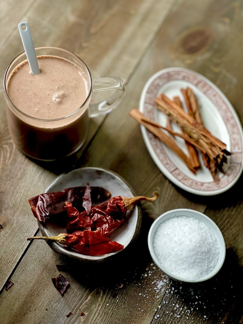 A clear glass mug of spiked hot chocolate with a small plate of dried red ancho chilis, a small oval dish of cinnamon sticks, and a small bowl of salt on a wooden surface