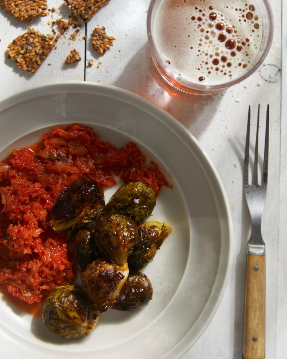 A white dish on a white surface with red kimchi and roasted Brussels sprouts with a serving fork, a cup of beer, and some broken seeded crackers above the bowl.