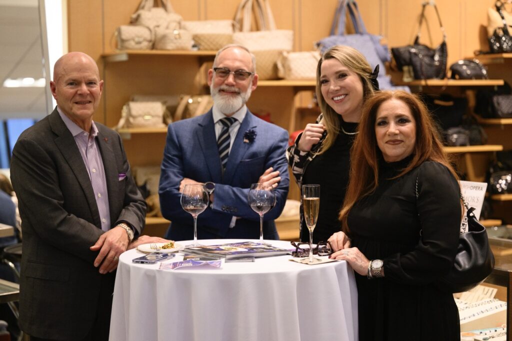 A group of four people stand at a white table with wine on it and purses behind them.
