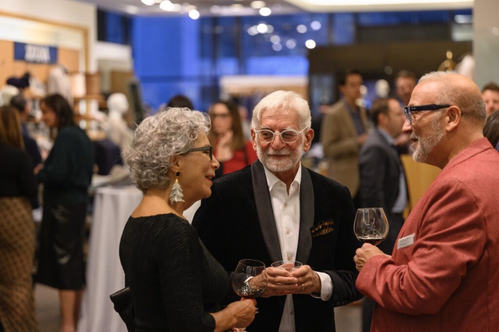 A woman in black stands talking to a man in black and a man in red with wine glasses in their hands.