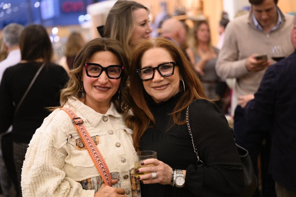 Two women with glasses stand together, one in white and one in black.