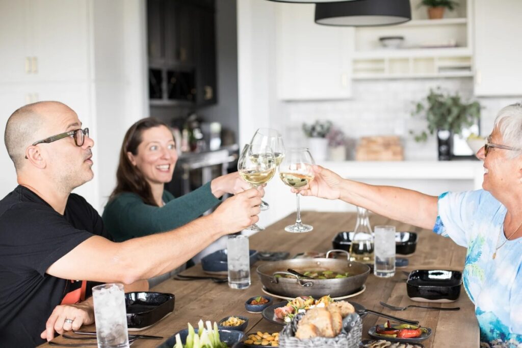 Justin Severino, his wife Hilary Prescott Severino, and an older woman toast with glasses of white wine at a brightly lit dinner table set with a paella pan and plates of charcuterie and fresh vegetables.