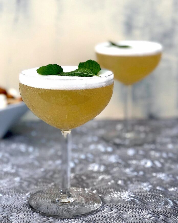 Keeping It Simple TABLE Magazine Two coupe glasses filled with a light yellow, frothy cocktail garnished with fresh mint leaves, likely featuring vodka and The Lemongrass ROOT Crafted Cocktail Mixer.