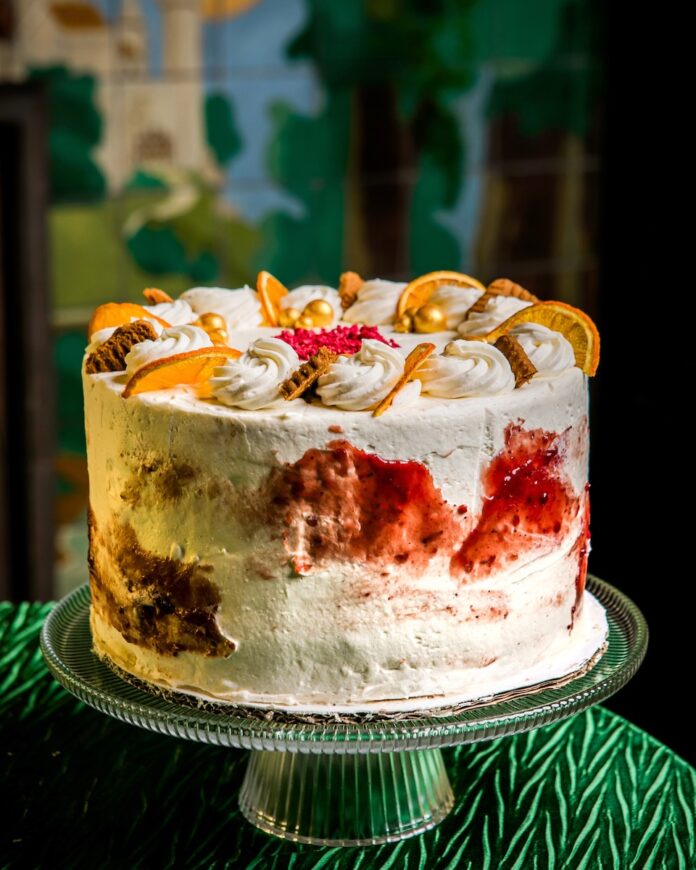 German Christmas Market in a Cake A cake with white icing decorated with orange slices and cranberry displayed on a glass cake plate on a table with a green table cloth.