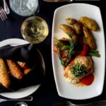 A plate of LeMont's Chicken Christina with roasted potatoes, asparagus, and a tomato basil coulis, served alongside bread, a glass of white wine, and water on a black tablecloth, emphasizing fine dining cuisine.