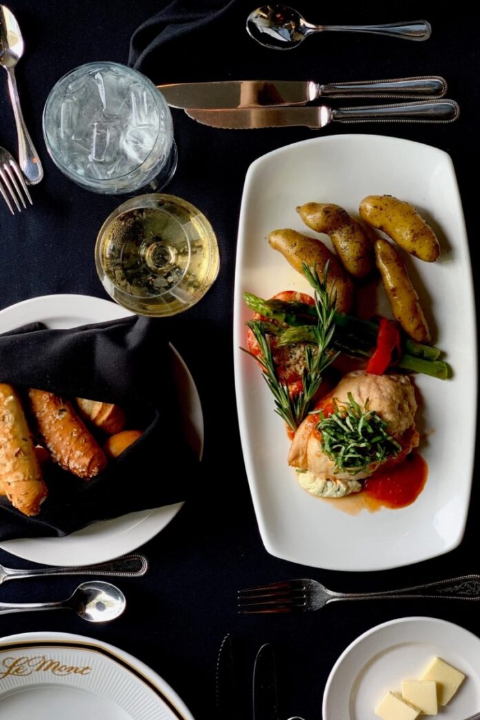A plate of LeMont's Chicken Christina with roasted potatoes, asparagus, and a tomato basil coulis, served alongside bread, a glass of white wine, and water on a black tablecloth, emphasizing fine dining cuisine.
