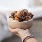 A white bowl full of Oliebollen Dutch donuts being held by a person as another sprinkles powdered sugar overtop.