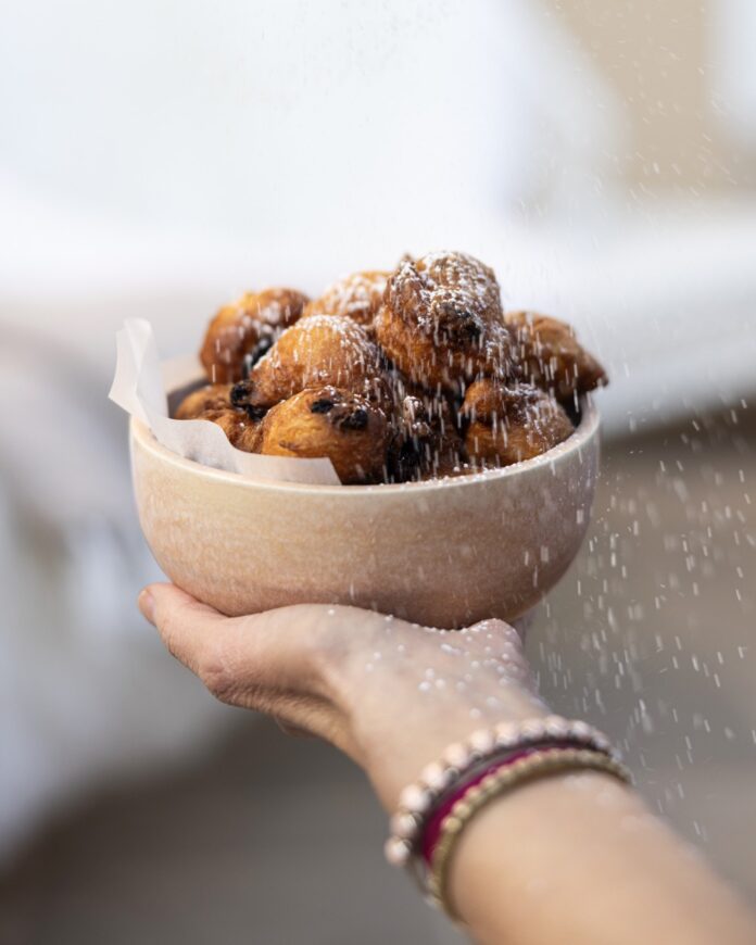 Oliebollen, Dutch Oil Ball Donuts A white bowl full of Oliebollen Dutch donuts being held by a person as another sprinkles powdered sugar overtop.