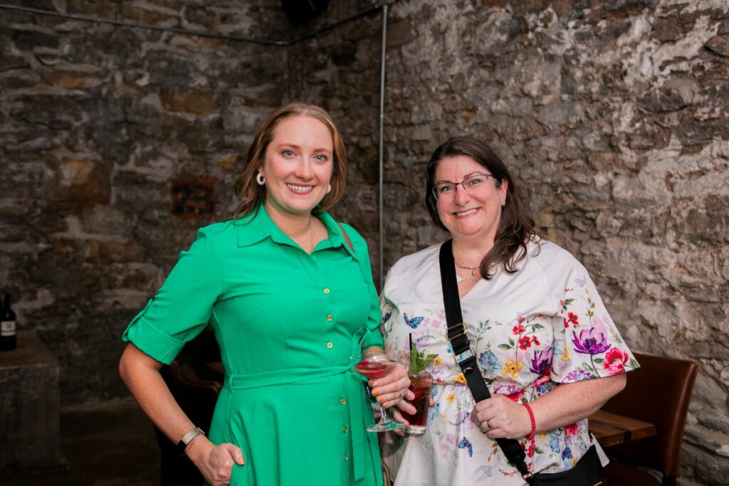 A smiling Nicole Harrell in a green dress and a friend in a floral blouse holding drinks, posing against a rustic stone wall at a non-profit networking mixer.
