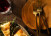 Thanksgiving Finger Foods Triangle cuts of a phyllo pastry with fresh herbs as garnish in between, on a round plate in the left corner, and a stack of terra cotta colored plates with three gold forks on a wooden surface.