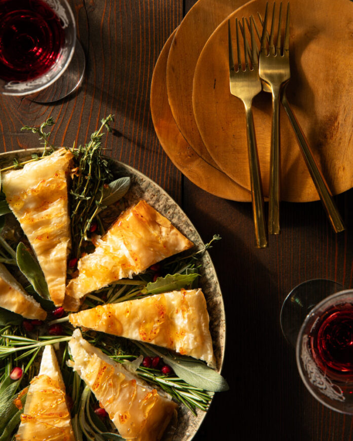 Pear and Goat Cheese Bites Triangle cuts of a phyllo pastry with fresh herbs as garnish in between, on a round plate in the left corner, and a stack of terra cotta colored plates with three gold forks on a wooden surface.