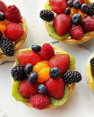Individual fruit tarts with custard filling, topped with bright strawberries, blueberries, blackberries, raspberries, and slices of kiwi.