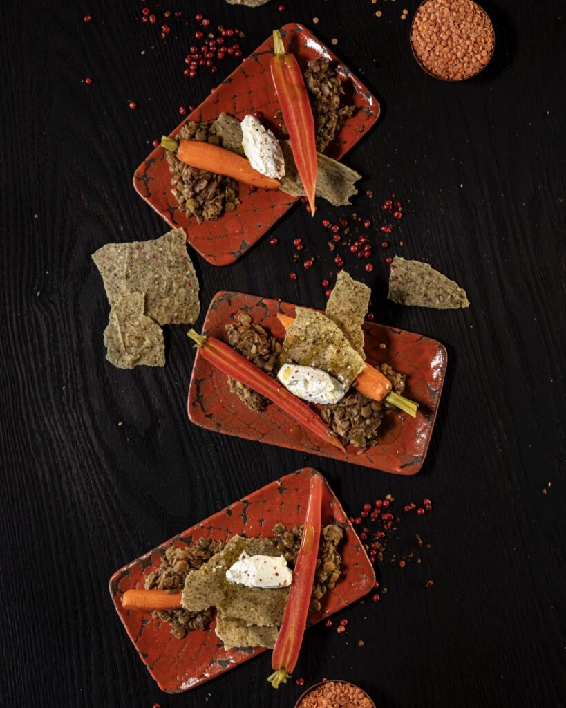 Three orange rectangular plates with pickled baby carrots, smoked labneh, and lentils.