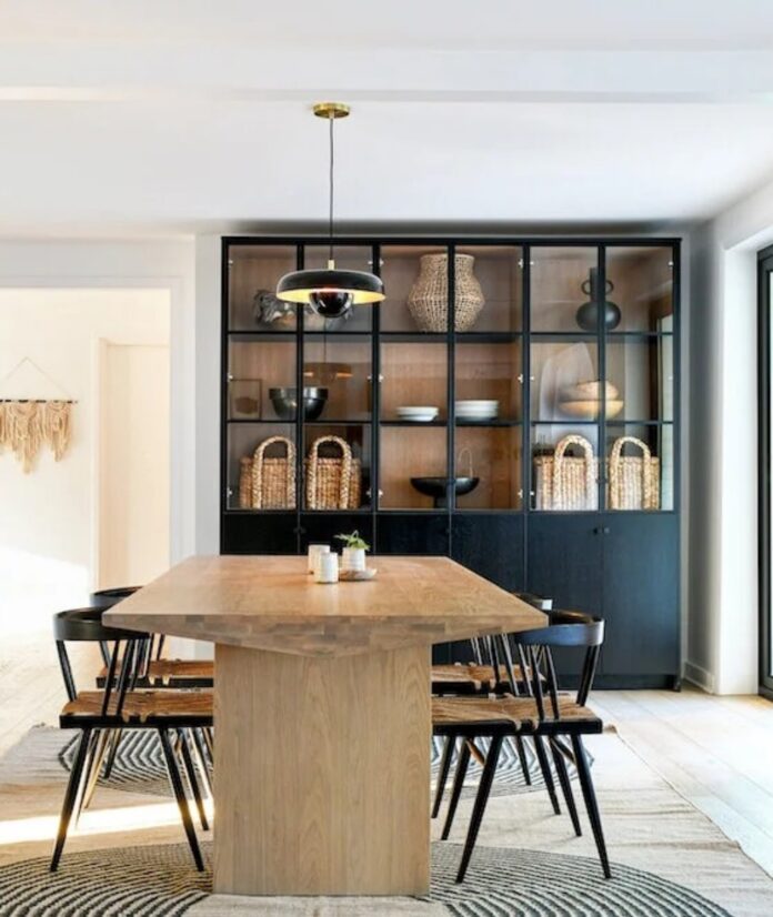 A modern dining room with a long natural wood table, black chairs with woven seats, a black display cabinet with woven baskets, and a hanging black and brass light fixture.